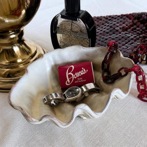 Puede incluir: Un plato de cerámica color crema con forma de concha que contiene un reloj de pulsera plateado y una pequeña caja de cerillas rectangular roja con el texto blanco Bone's Steak and Seafood, acompañado de una cadena de plástico roja.