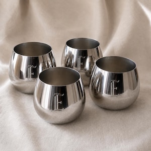 Op de afbeelding: Een set van vier afgeronde, gepolijste zilverkleurige metalen bekers. Elke beker heeft een glad, reflecterend oppervlak en is aan de voorzijde gegraveerd met een enkele, sierlijke hoofdletter E in een klassiek gotisch lettertype.