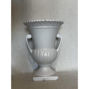 Può includere: Un'urna in ceramica grigio chiaro con due manici curvi e una base quadrata. L'urna presenta un design scanalato dettagliato attorno alla base e al bordo. La forma generale è simmetrica, con un'estetica classica e antica. Per scopi decorativi.