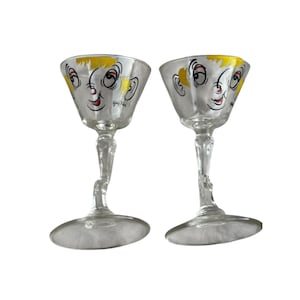 May include: Two clear stemmed cocktail glasses with a whimsical face design. The faces have yellow hair, red lips, and black eyes. The glasses have a unique, curved stem and a wide, shallow bowl.
