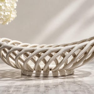 May include: An oval-shaped ceramic bowl featuring a cream-colored, open-weave lattice design. The structure consists of interconnected, curved ceramic strips that form a basket-like pattern, resting on a solid, circular base with a smooth finish.