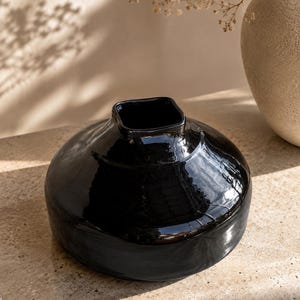May include: A low-profile, wide-bodied ceramic vase with a glossy black finish. The vessel features a rounded, disc-like shape that tapers upward to a small, square-shaped opening at the top, creating a distinct contrast between the curves and angles.