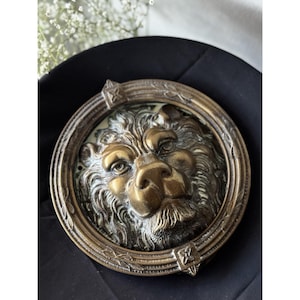 May include: Antique brass lion door knocker. The knocker features a detailed lion's head within a circular frame. The lion's face has intricate detailing, with a prominent nose and expressive eyes. The frame has decorative elements. The overall color is a warm, aged brass.