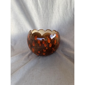 May include: A round ceramic bowl with a scalloped rim. The bowl is brown with orange polka dots and a cream-colored interior. The bowl is likely handmade and suitable for decorative use.