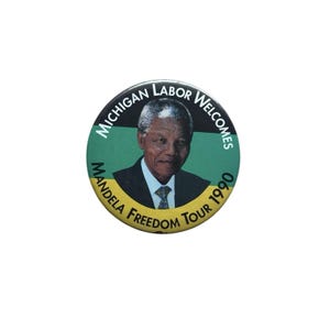 May include: A round button with a portrait of Nelson Mandela, with the text "Michigan Labor Welcomes Mandela Freedom Tour 1990" in a circular design. The button has a green and black background.
