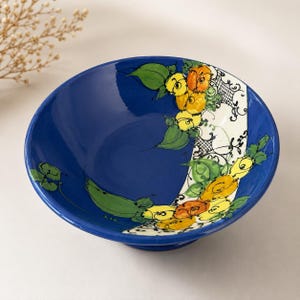 May include: A deep blue ceramic bowl featuring a hand-painted floral design with yellow and orange flowers, green leaves, and black filigree accents on a white curved band that sweeps across the interior surface of the bowl.
