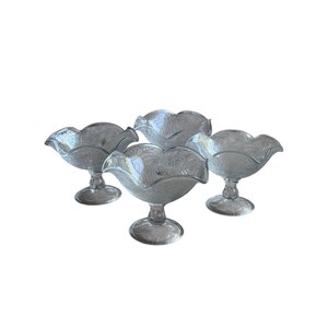 May include: Four clear glass dessert bowls with a floral pattern and scalloped edges. The bowls are elevated on short, decorative stems. The glass has a slight grey tint, and the bowls are arranged on a white background.