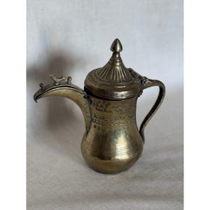 Puede incluir: Una cafetera de latón vintage con un pico curvo, asa y tapa decorativa. La cafetera presenta patrones grabados y una pequeña figura de pájaro en el pico. El diseño sugiere un estilo antiguo o de Oriente Medio.