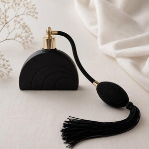 May include: A matte black perfume bottle shaped like a semi-circle with concentric arch detailing. The bottle features a gold-toned metal collar and a black fabric-covered atomizer bulb connected by a tube, finished with a long black tassel.