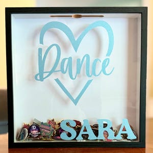Puede incluir: Una caja de sombra con marco negro, con un corazón azul claro y la palabra "Dance" en cursiva. El nombre "SARA" se muestra debajo, con varios alfileres y baratijas en el interior. El fondo es blanco.
