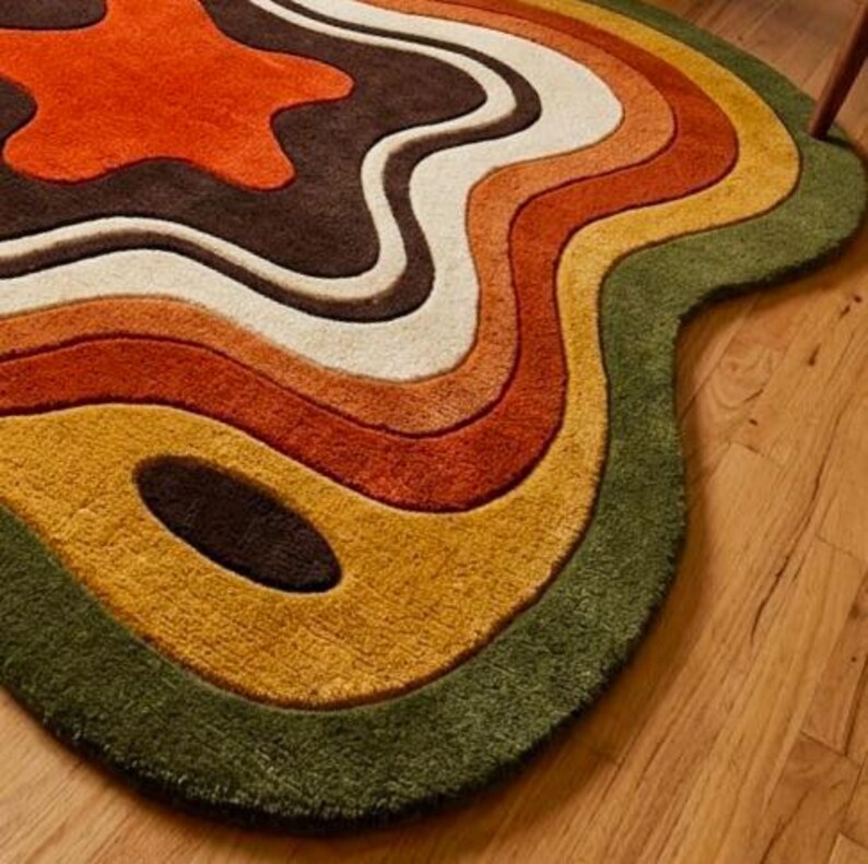 Puede incluir: Una alfombra con una forma org&aacute;nica irregular y bandas de colores onduladas y conc&eacute;ntricas. La paleta incluye tonos de verde bosque profundo, amarillo mostaza, naranja quemado, crema y marr&oacute;n oscuro, creando un efecto topogr&aacute;fico.
