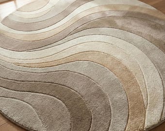 May include: A circular area rug featuring a series of flowing, wave-like bands in varying shades of beige, cream, tan, and light brown. The rug has a textured, tufted pile surface with curved lines that create a rhythmic, layered visual pattern.