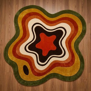 Puede incluir: Una alfombra de forma irregular similar a una ameba con capas conc&eacute;ntricas y onduladas. La paleta incluye tonos verde oscuro, amarillo mostaza, naranja quemado, crema y marr&oacute;n oscuro, con una forma de estrella naranja en el centro.