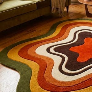 Puede incluir: Una alfombra con una forma org&aacute;nica irregular y bandas de colores conc&eacute;ntricas y onduladas. La paleta incluye verde oliva intenso, amarillo mostaza, naranja quemado, crema y marr&oacute;n chocolate oscuro, creando un dise&ntilde;o fluido y en capas.