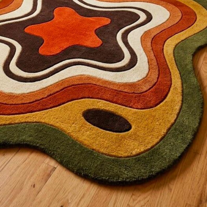Puede incluir: Una alfombra de forma irregular y org&aacute;nica con bandas de color conc&eacute;ntricas y onduladas. La paleta incluye tonos de naranja brillante, marr&oacute;n oscuro, crema, amarillo mostaza y verde oliva, creando un aspecto topogr&aacute;fico en capas.