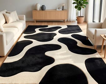 Peut inclure: Un tapis rectangulaire présentant un motif abstrait contrasté rappelant une peau de vache, avec de grandes formes organiques noires sur un fond crème uni. Le tapis possède un poil dense et texturé, recouvrant le sol entre deux canapés.
