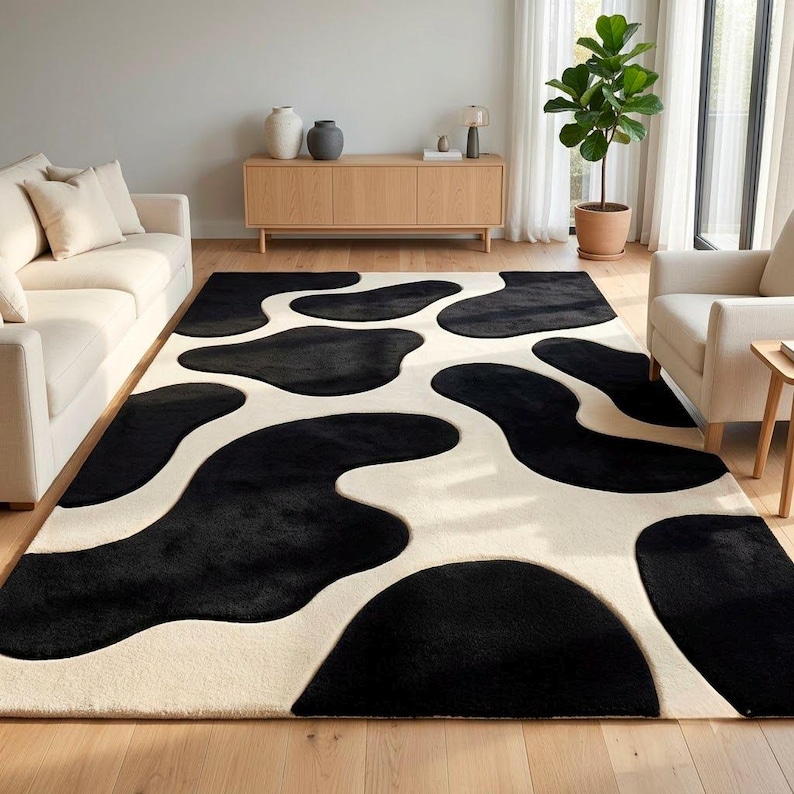 May include: A rectangular area rug featuring a high-contrast, abstract cow-print pattern with large, organic black shapes set against a solid cream-colored background. The rug has a plush, textured pile and covers the floor between two sofas.