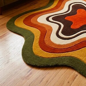 Puede incluir: Una alfombra de forma org&aacute;nica irregular con bandas de color conc&eacute;ntricas y onduladas. La paleta incluye verde oliva oscuro, amarillo mostaza, naranja quemado, crema y marr&oacute;n chocolate oscuro, creando una apariencia fluida y texturizada.