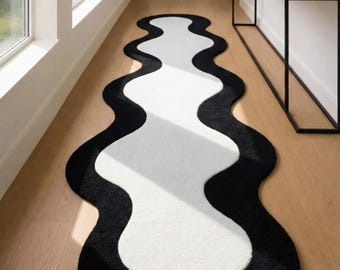 May include: A modern, abstract rug with a wavy design. The rug features a white center bordered by a thick, black edge. The rug is placed on a light-colored wooden floor in a hallway.