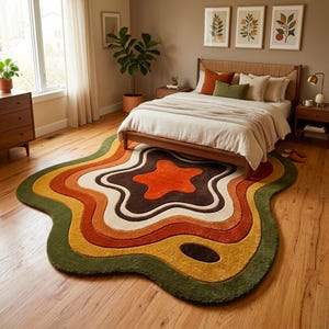 Puede incluir: Una alfombra de forma org&aacute;nica irregular con capas conc&eacute;ntricas onduladas en tonos verde oliva, amarillo mostaza, naranja quemado, marr&oacute;n y crema. La alfombra tiene un pelo grueso y texturizado y est&aacute; colocada sobre un suelo de madera clara.
