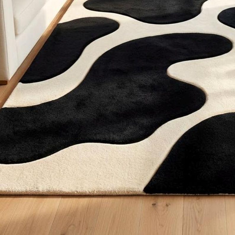 May include: A plush area rug featuring a high-contrast pattern of large, irregular black organic shapes set against a solid cream-colored background. The rug has a soft, textured pile surface that creates a distinct visual depth between the colors.