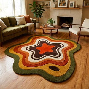 Puede incluir: Una alfombra de forma org&aacute;nica irregular con bandas conc&eacute;ntricas onduladas en colores verde oliva oscuro, amarillo mostaza, naranja quemado, crema y marr&oacute;n oscuro. La alfombra tiene un pelo texturizado y denso sobre un suelo de madera clara.