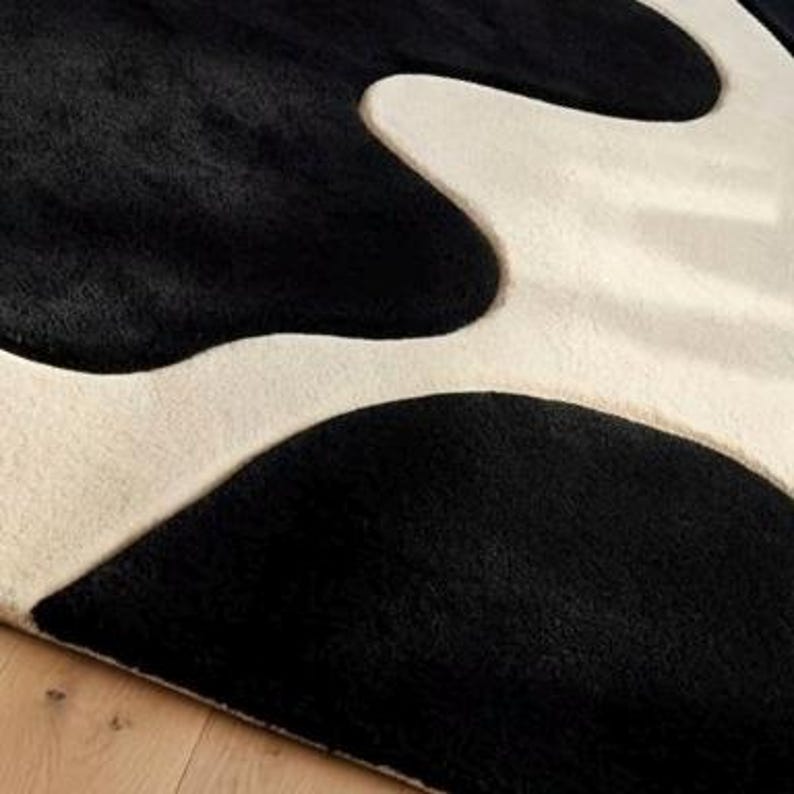May include: A plush area rug featuring a high-contrast pattern of large, organic black shapes set against a creamy off-white background. The surface has a dense, soft-textured pile with clean, curved edges defining the abstract color blocks.