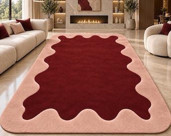Peut inclure: Un tapis rectangulaire présentant un centre bordeaux profond avec une bordure aux bords ondulés et festonnés dans un rose poudré doux. Le tapis possède une texture pelucheuse de couleur unie et des coins arrondis, créant un contraste net.