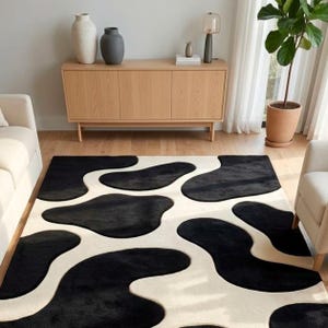 May include: A rectangular area rug featuring a high-contrast pattern of large, irregular black organic shapes set against a solid cream-colored background. The rug has a plush, textured surface and covers a significant portion of the floor space.
