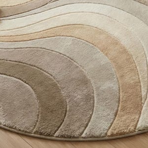 May include: A round rug with a wave pattern in shades of beige, tan, and brown. The rug has a soft, textured surface and a neutral color palette, suitable for various interior styles. The design features curved lines.
