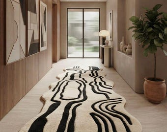 Peut inclure: Un tapis de forme organique et irrégulière présentant une base de couleur crème avec des motifs de lignes noires fluides et audacieuses. Le tapis est placé au centre sur un sol clair, s'étendant le long d'un couloir aux murs lambrissés.
