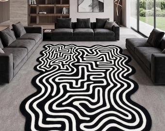 Peut inclure: Un grand tapis de sol présentant une forme organique irrégulière avec un motif contrasté noir et blanc. Le design se compose de lignes épaisses, continues et sinueuses qui créent un effet de labyrinthe sur toute la surface du revêtement.