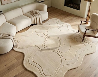 May include: Beige, abstract-shaped area rug with a raised, wavy pattern. The rug is placed on a hardwood floor in a living room setting, alongside a cream-colored sofa and a mid-century modern chair.