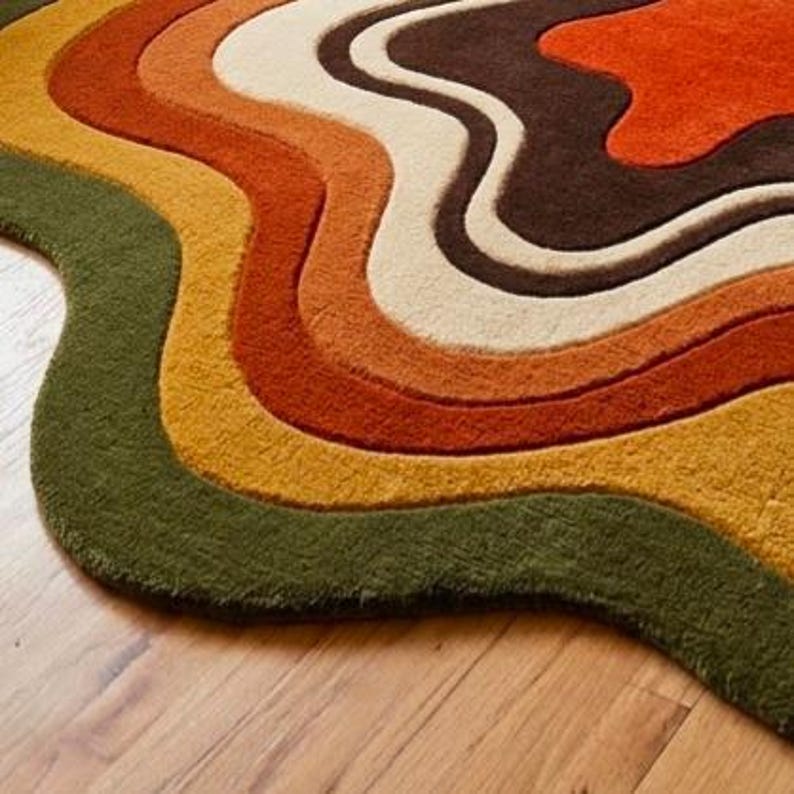 Puede incluir: Una alfombra de pelo insertado que presenta una serie de bandas onduladas y org&aacute;nicas en tonos verde oliva, amarillo mostaza, crema, naranja quemado y marr&oacute;n oscuro. La alfombra tiene un pelo grueso y texturizado con una forma de contorno libre.