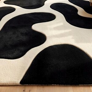 May include: A plush area rug featuring a high-contrast pattern of large, irregular black organic shapes set against a solid cream-colored background. The rug has a thick, soft-textured pile with distinct, rounded edges defining the dark patches.