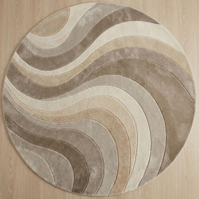 May include: A round rug with a wave pattern in shades of beige, tan, and brown. The rug's design features curved lines that create a flowing, textured appearance. The rug is placed on a light-colored wooden floor.