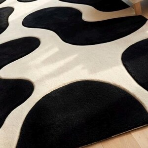 May include: A plush area rug featuring a high-contrast pattern of large, irregular black organic shapes set against a creamy off-white background. The surface has a soft, textured pile with distinct curved edges defining the black and white sections.