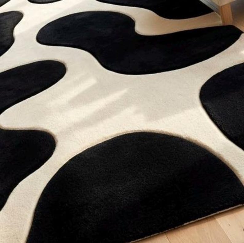 May include: A plush area rug featuring a high-contrast pattern of large, irregular black organic shapes set against a creamy off-white background. The surface has a soft, textured pile with distinct curved edges defining the black and white sections.