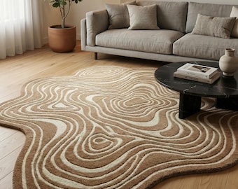 Puede incluir: Una alfombra de área marrón y crema con una forma orgánica única. La alfombra presenta un diseño topográfico en capas con líneas arremolinadas. La alfombra está colocada sobre un piso de madera, cerca de un sofá gris y una mesa de centro negra.