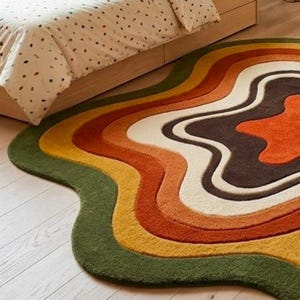 Puede incluir: Una alfombra con forma irregular y ondulada que presenta bandas conc&eacute;ntricas de colores fluidos. La paleta incluye verde oliva oscuro, amarillo mostaza, naranja quemado, crema y marr&oacute;n oscuro, creando un aspecto estratificado.