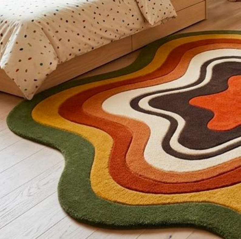 Puede incluir: Una alfombra con forma irregular y ondulada que presenta bandas conc&eacute;ntricas de colores fluidos. La paleta incluye verde oliva oscuro, amarillo mostaza, naranja quemado, crema y marr&oacute;n oscuro, creando un aspecto estratificado.