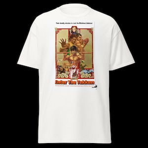 Puede incluir: Camiseta blanca con un gráfico colorido de "Enter The Tekken". El diseño incluye figuras de acción, el texto "Enter The Tekken" y la frase "Their deadly mission to crack the Mishima Zaibatsu!"