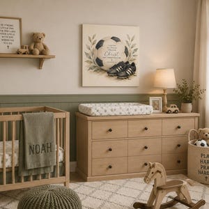 May include: A square wall canvas featuring a soccer ball and black cleats with the text I CAN DO ALL THINGS THROUGH CHRIST WHO STRENGTHENS ME. The artwork is displayed in a nursery room with a wooden crib, a dresser, and various toys.