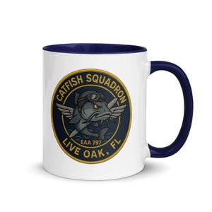 May include: White ceramic mug with a navy blue interior and handle. The mug features a circular emblem with a catfish wearing goggles and wings, the text "CATFISH SQUADRON" and "LIVE OAK, FL".
