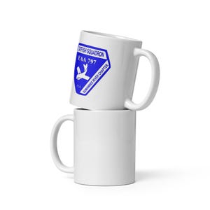May include: Two white ceramic mugs stacked on top of each other. The top mug features a blue emblem with the text "Catfish Squadron EAA 797 Suwannee River Chapter FLA" and a graphic of two stylized airplanes. The bottom mug is plain white.