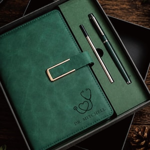 May include: A dark green notebook with a gold-toned metal clasp and a stethoscope heart icon engraved above the text DR. MITCHELL. The set includes a matching dark green pen and a silver ink refill, all presented in a dark gray gift box.