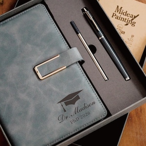 Può includere: Un set regalo con un diario in similpelle grigio con chiusura dorata, una penna e una ricarica. Il diario è inciso con un cappello di laurea e il testo "Dr. Madison, PhD 2026". È incluso anche un piccolo blocco note con il testo "Mideav Painting".