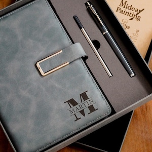 Puede incluir: Un juego de regalo que incluye un diario de cuero gris con cierre dorado, personalizado con el nombre "MARTIN". También incluye un bolígrafo negro, un bolígrafo plateado y un pequeño bloc de notas con el texto "Midea Painting".