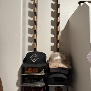 May include: A wooden hat rack mounted on a wall, holding several baseball caps. The caps are stacked and feature various designs, including an American flag and the text "SALTY CREW". The rack is made of dark wood.