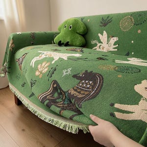 May include: Green sofa throw featuring a repeating pattern of animals and botanical designs. The throw has a fringe trim and is draped over a wooden-framed sofa. A green plush toy sits on the sofa. The throw is made of woven fabric.